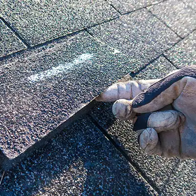 Worker's hand on a roof lifting up an asphalt shingle to inspect underneath it